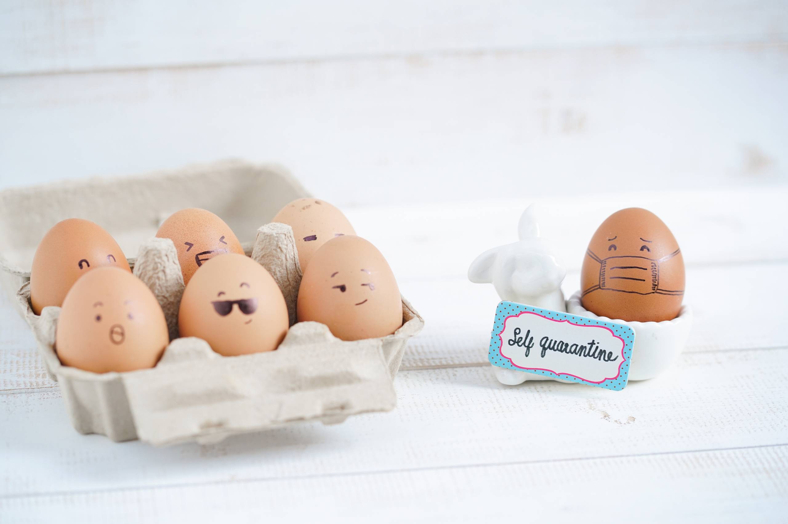 Six Eggs in a carton with sharpie drawn faces and one egg off to the side with a sharpie drawn mask and a sign saying "self quarantine"
