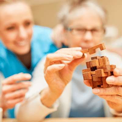 Woman in memory care facility putting wooden puzzle together while female nurse helps
