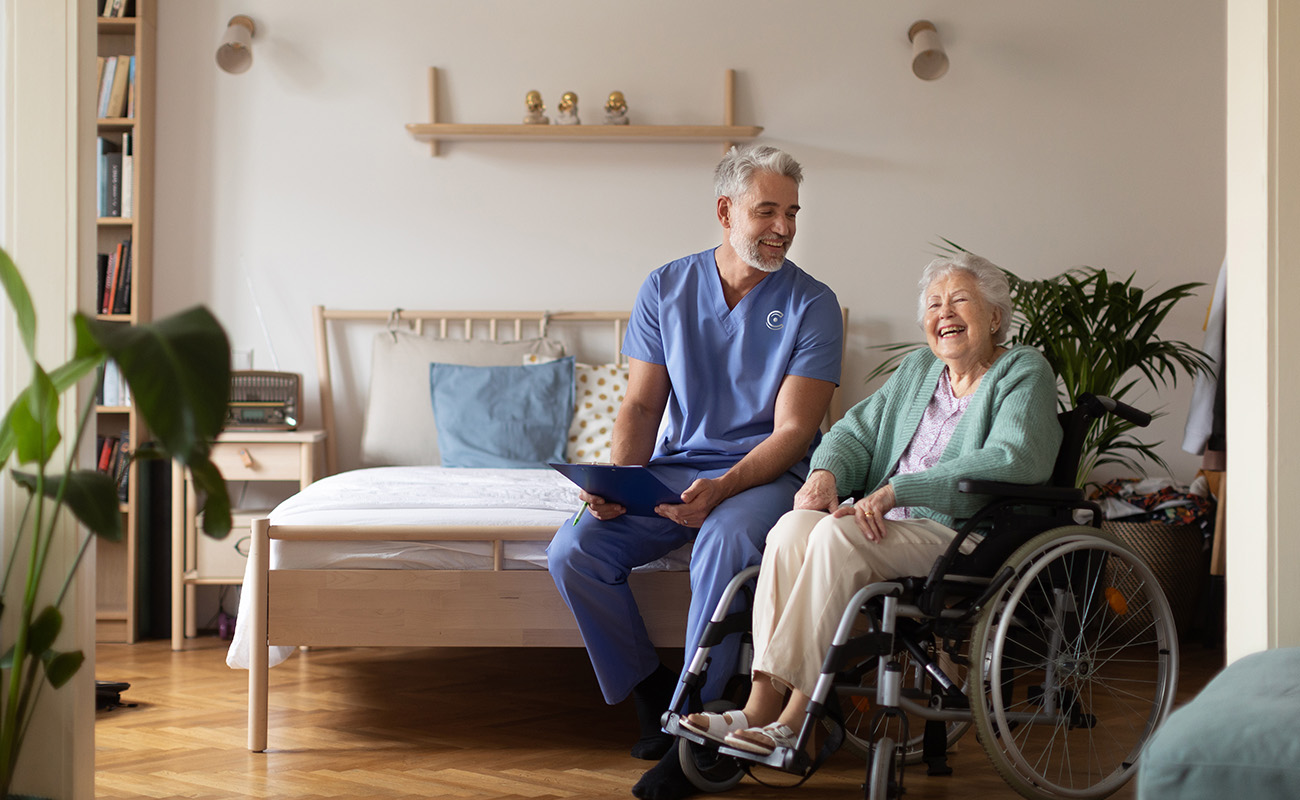 Caregiver in blue uniform smiling with senior woman in wheelchair in cozy living unit.