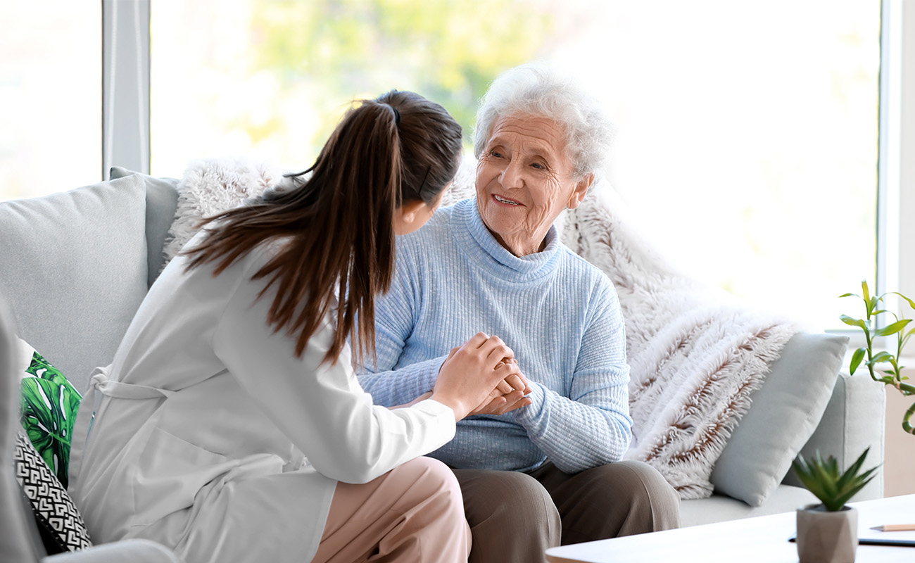 Smiling elderly woman in blue sweater speaks to a caregiver indoors.