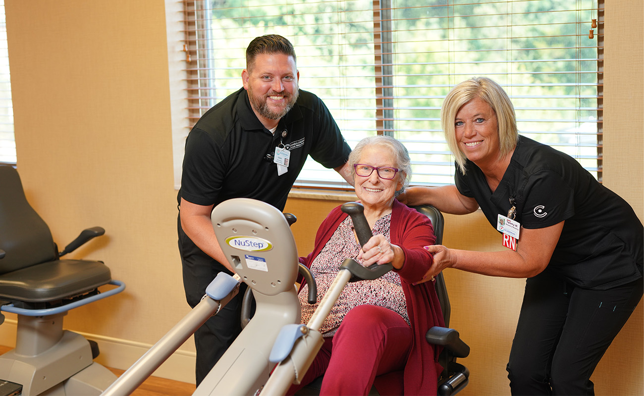Senior woman using exercise equipment with help from caregivers in a community room.