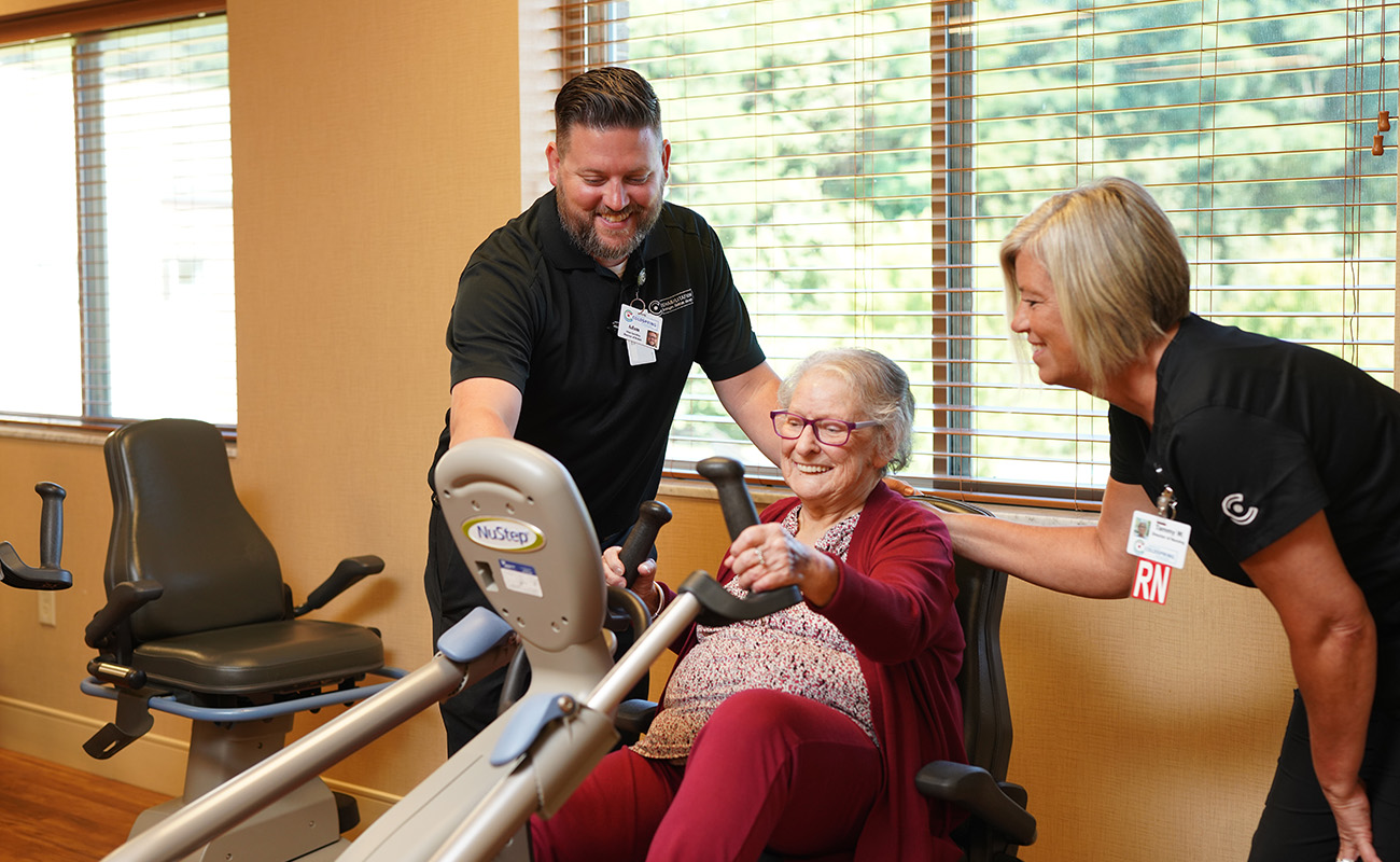 Elderly woman on exercise machine assisted by staff in a bright room.