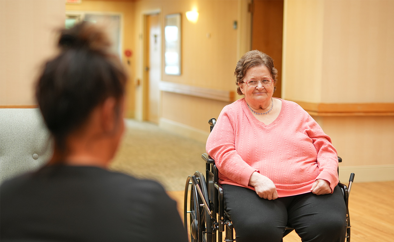 Woman in a wheelchair in a senior living community hallway next to another seated person.
