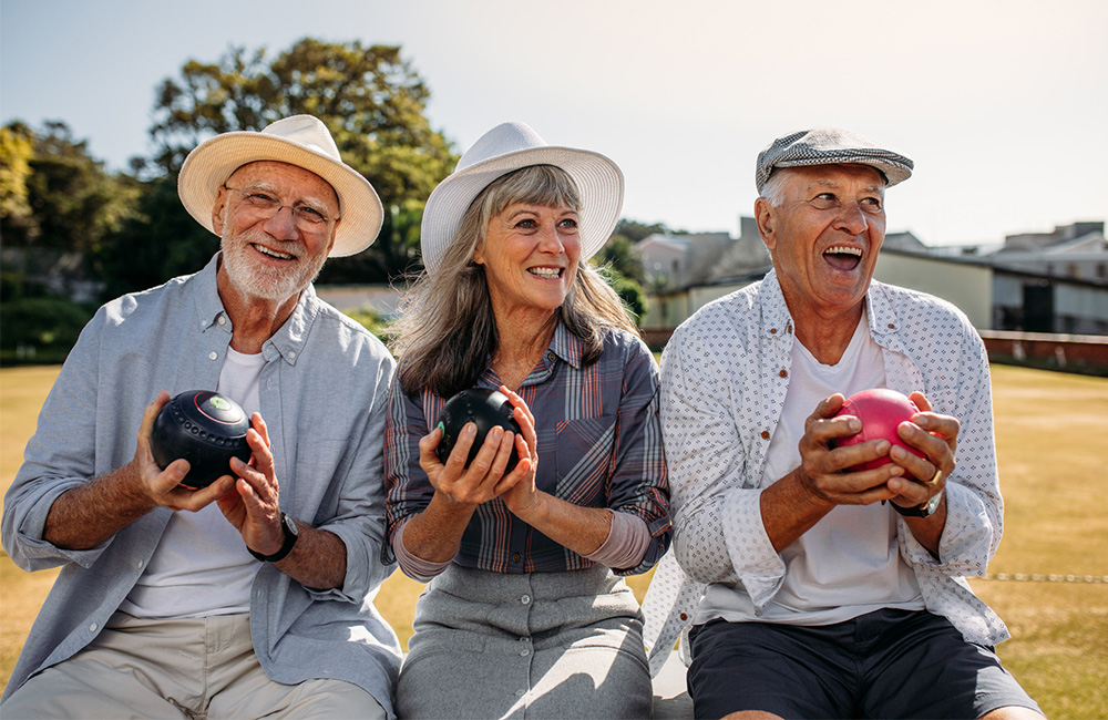 Three seniors playing bocce ball outdoors in bright, sunny weather, smiling and enjoying the game.