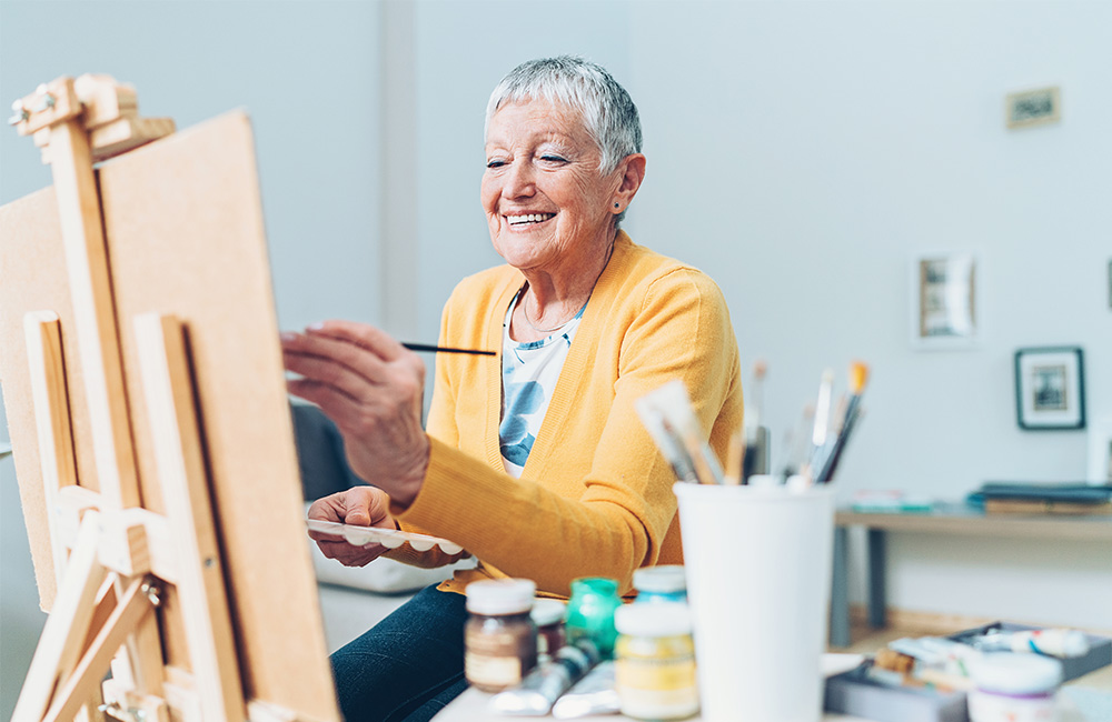 Smiling older adult painting on canvas in a cozy room with art supplies.