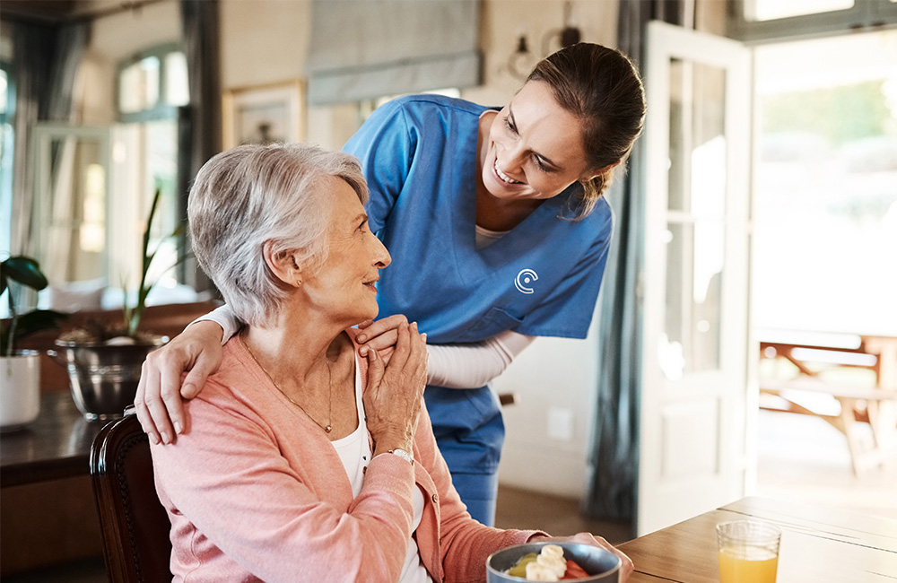 Smiling caregiver in blue uniform assists a senior woman in a pink sweater at a table.
