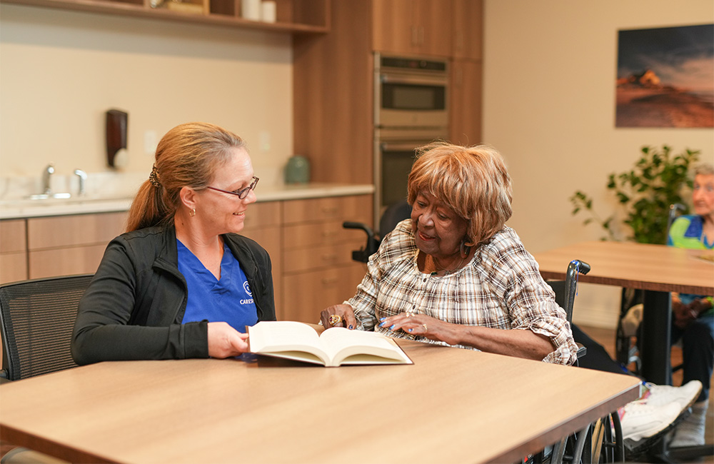 A caregiver reading a book with an elderly woman in a senior living unit common area.