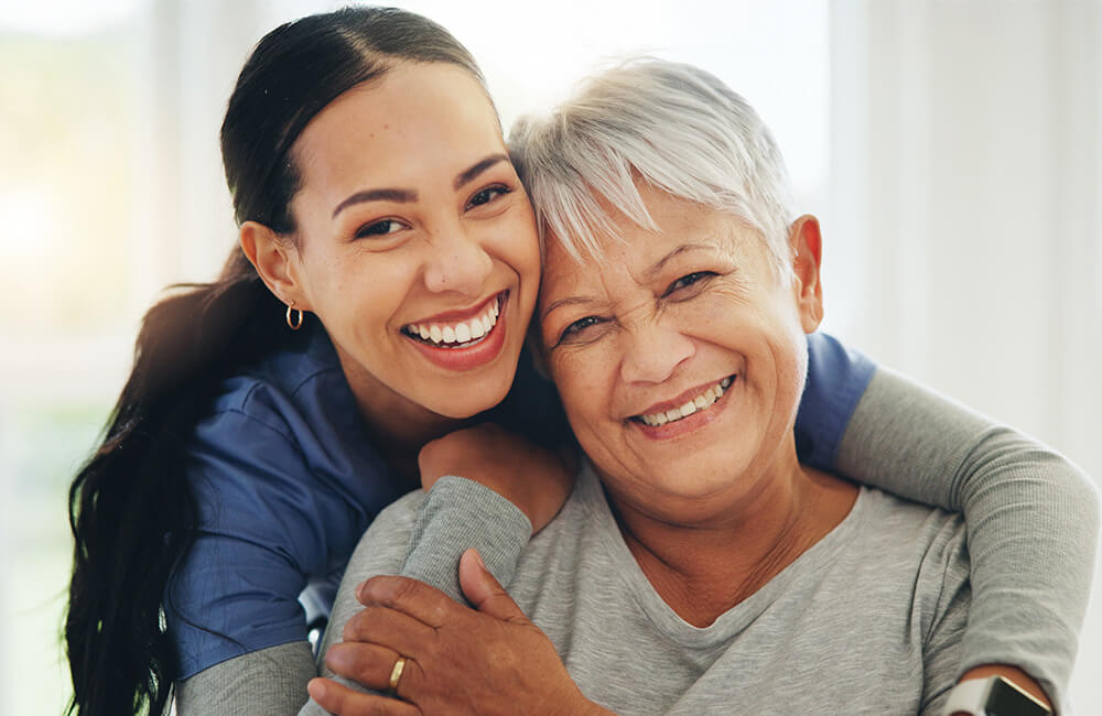 Young caregiver embraces smiling senior woman indoors.