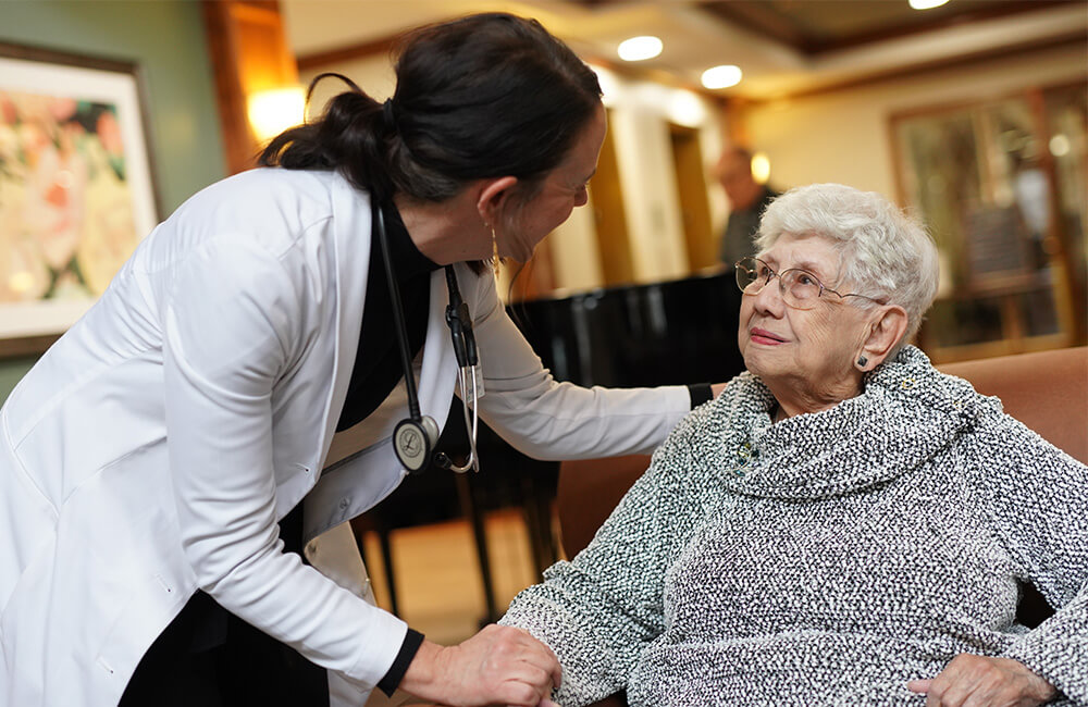 Caregiver supports elderly woman in senior community living room.