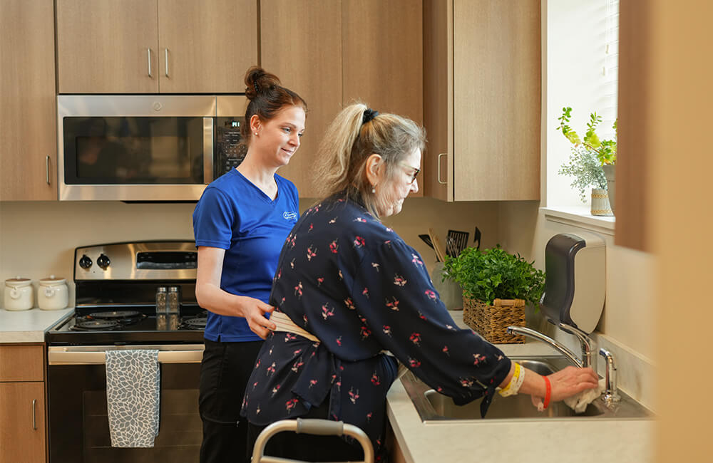 Caregiver assists woman at kitchen sink in a senior living unit.