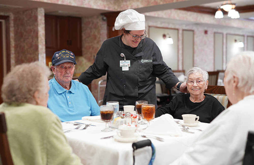 Chef interacting with seniors seated around a dining table with drinks and tableware.