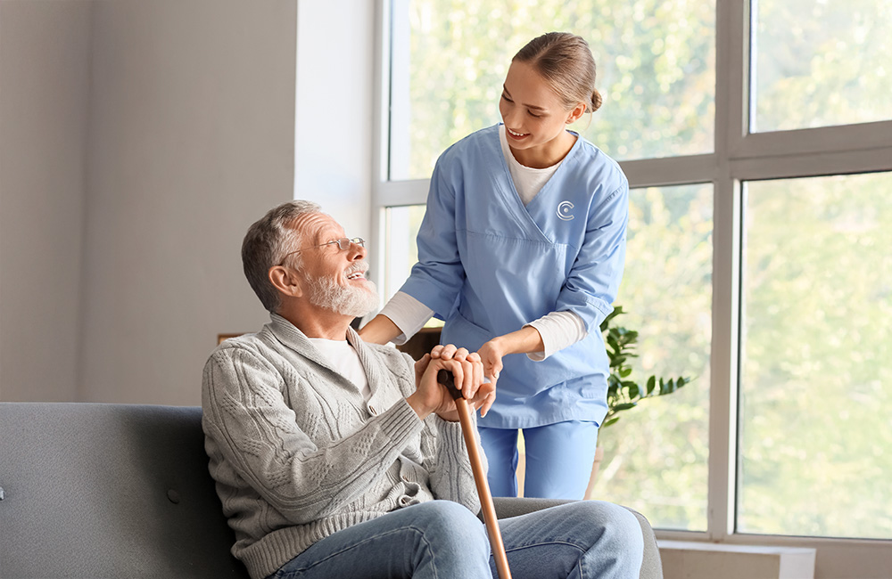 Caregiver assists senior man with a cane in a well-lit community space.