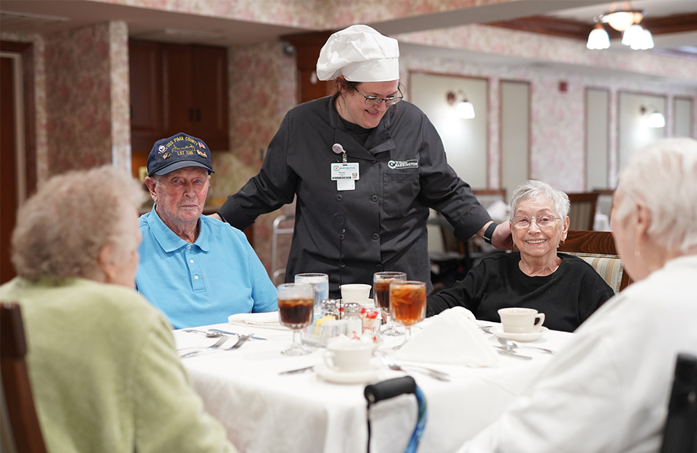 Chef conversing with seniors seated around a dining table in a communal dining area.