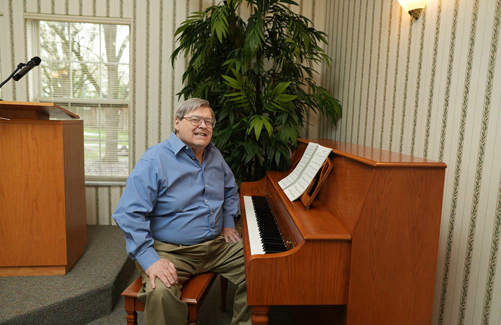 Smiling man in blue shirt sits at piano in community lounge with plant and podium nearby.