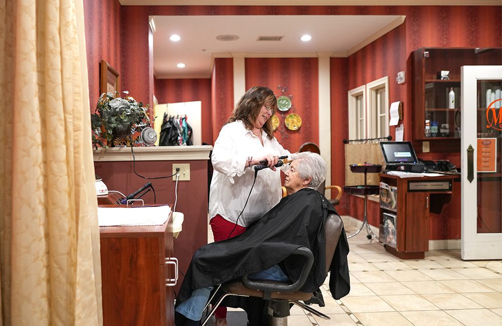 Hair stylist curling senior residents hair in community salon with red walls.