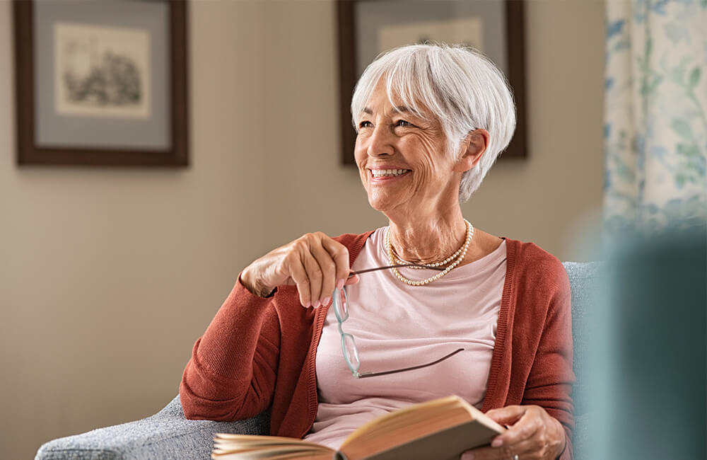 Elderly woman smiling while holding glasses and book in cozy living room.