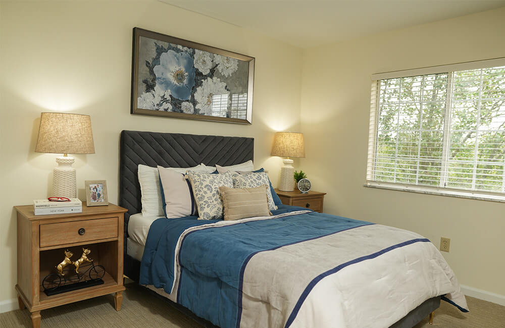 Cozy bedroom with twin beds, wooden nightstand, lamp, and flowers beneath a large window.
