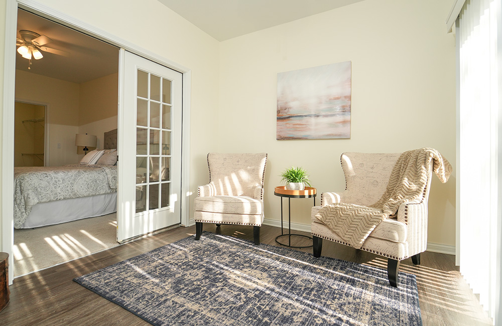 Bright senior living unit with chairs, table, and view of bedroom through glass doors.