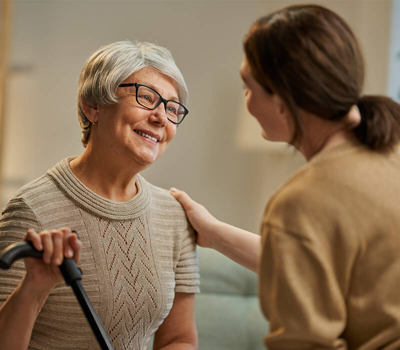 Elderly woman with glasses smiling while talking to a caregiver in a cozy room.