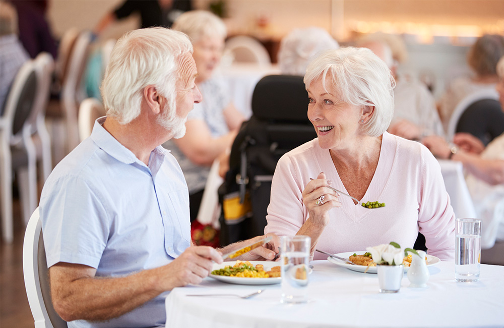 Elderly couple smiling and dining in a senior living community dining area.