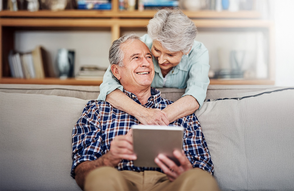 Elderly couple enjoying time together on a couch, woman embracing man holding a tablet.