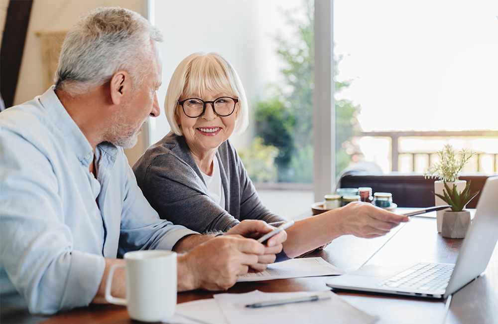 Smiling elderly couple using a laptop at a table in a bright living space.