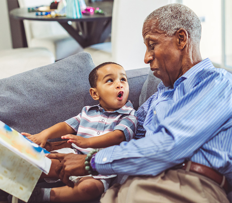 Elderly man reads a book to an excited child on a sofa in a living community.