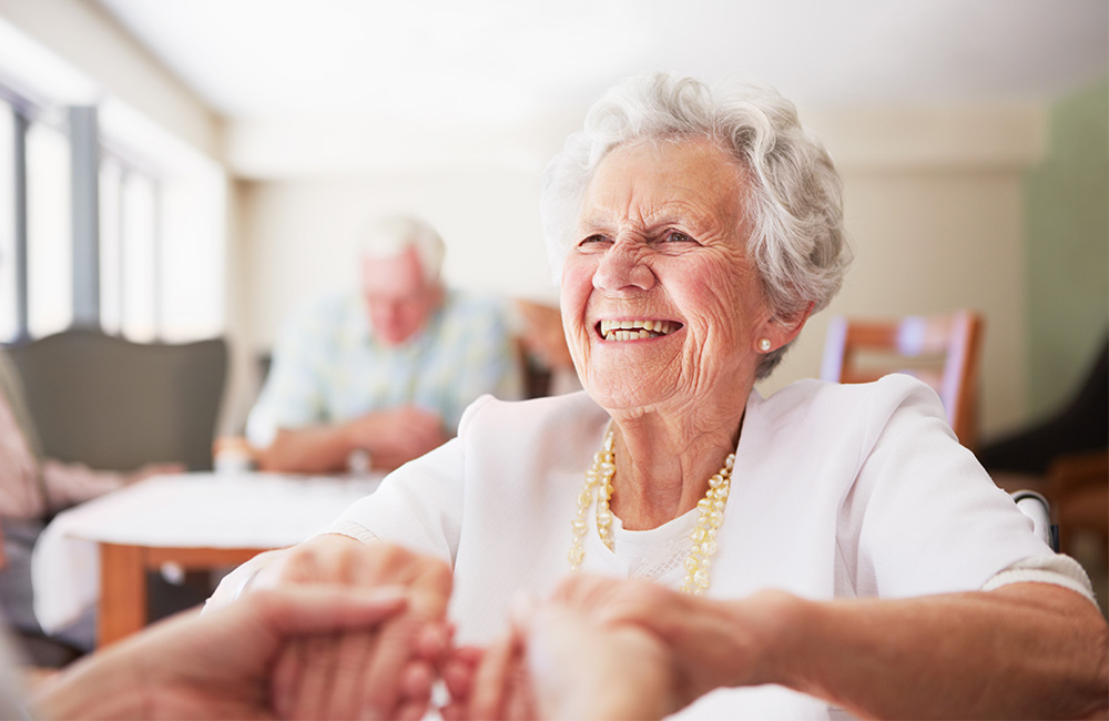 Smiling elderly woman holding hands in a bright community dining area.