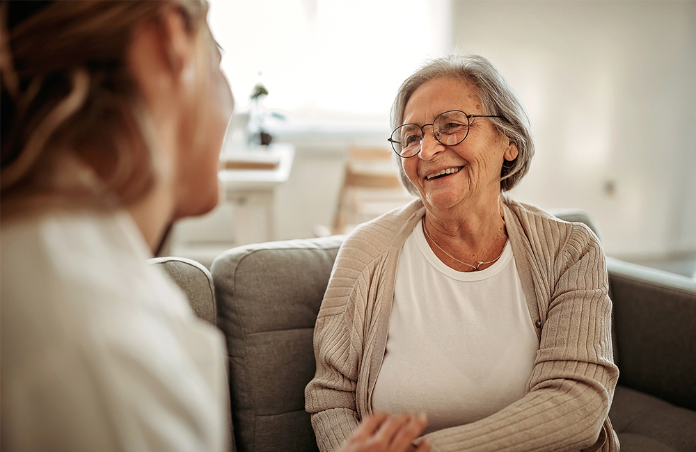 Elderly woman smiling and chatting with a caregiver in a cozy living room.