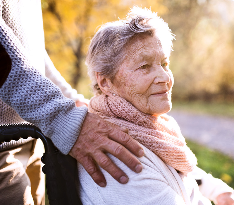 Elderly woman in a wheelchair with a comforting hand on her shoulder outdoors.