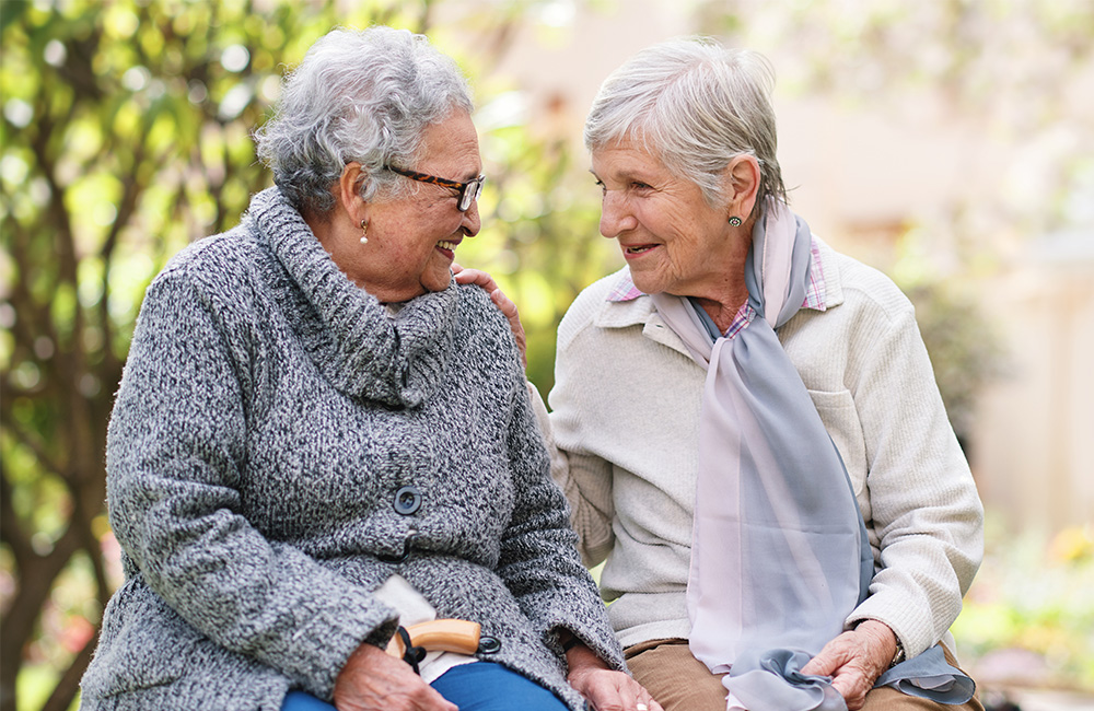 Two elderly women smiling at each other on a bench in a garden setting.