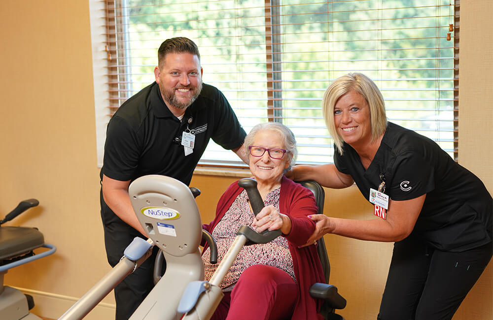 Senior resident using exercise equipment with assistance from two caregivers.
