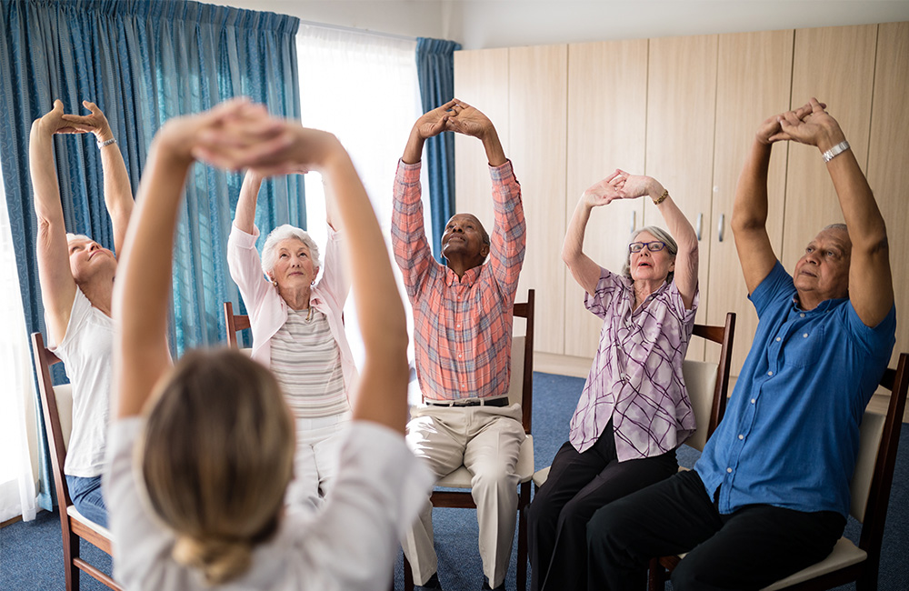 Group of seniors sitting indoors, participating in a seated stretching exercise.