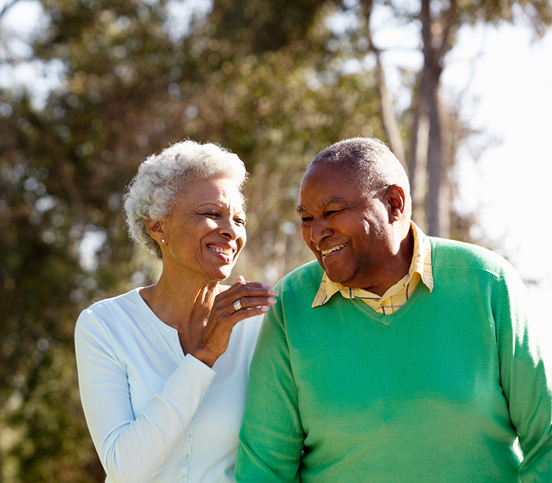 Elderly couple smiling and walking in a wooded area.