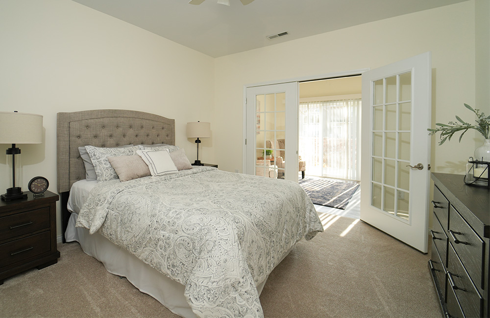 Spacious bedroom in a unit with light decor and large windows in a senior living community.