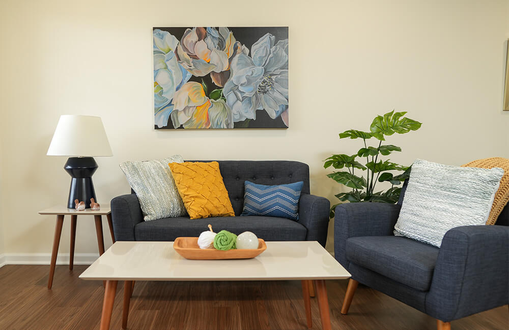Cozy unit living room with dark chairs, vibrant pillows, and floral painting.