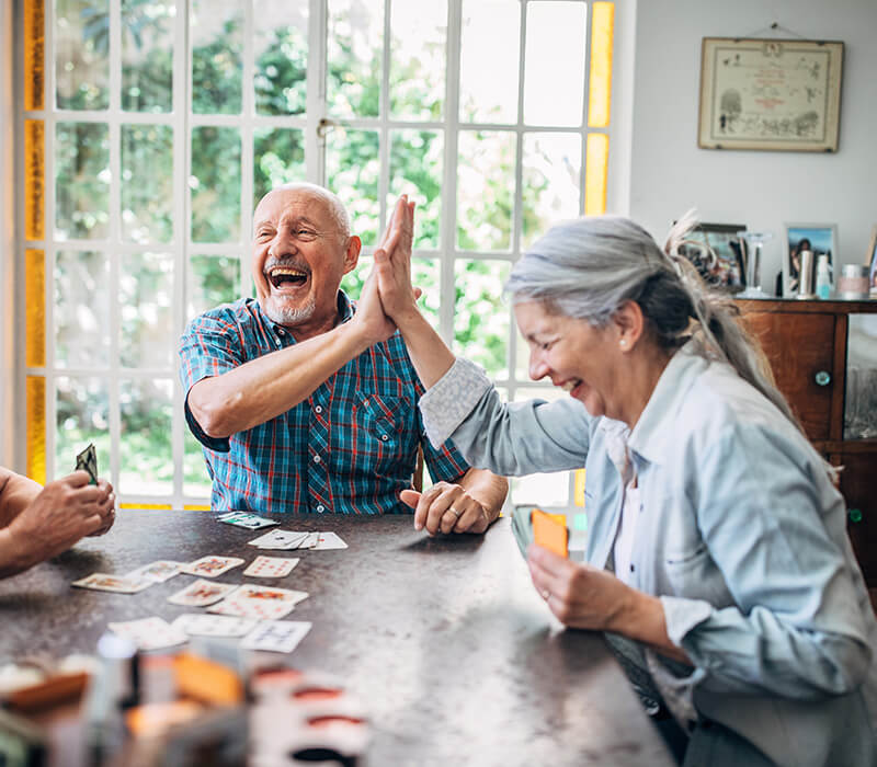 Elderly man and woman sharing a joyful high-five while playing cards indoors.
