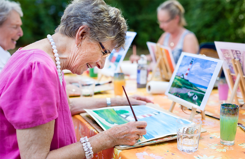 Elderly woman painting joyfully in outdoor art at community table.