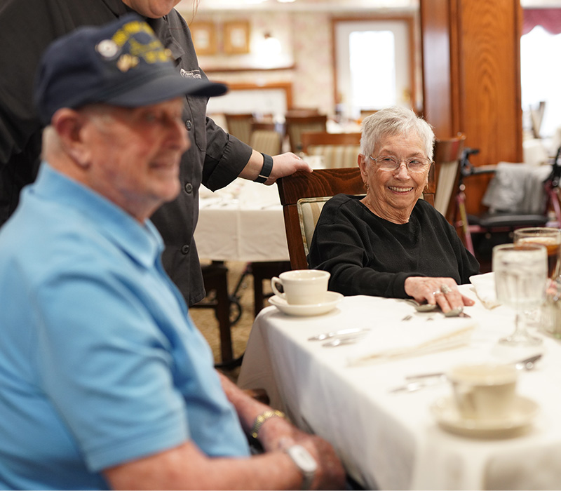 Two seniors dining at a table, one smiling with a caregivers hand on their shoulder.