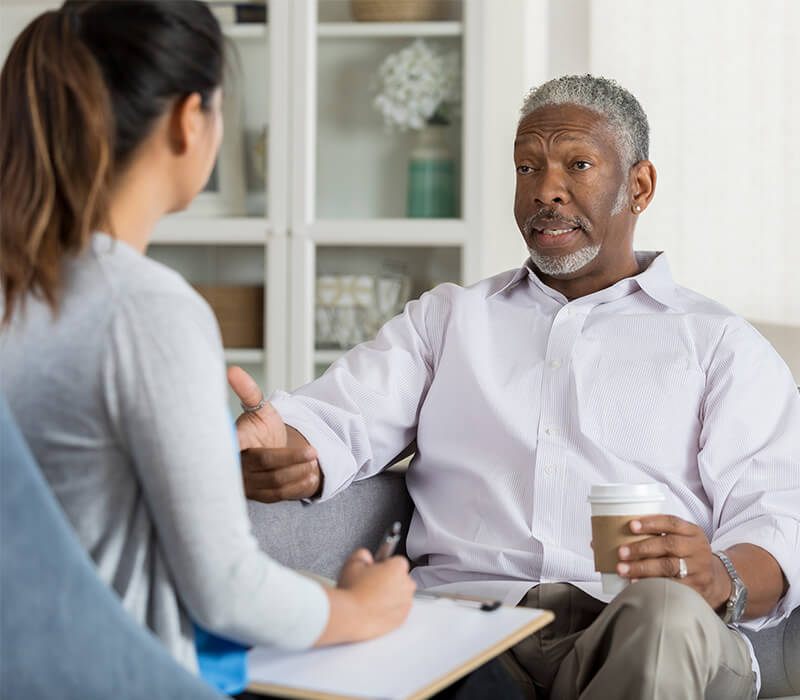 Elderly man holding coffee, engaged in conversation with a young woman taking notes.