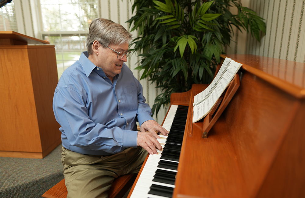 Senior man in blue shirt playing piano near large plant with open sheet music.
