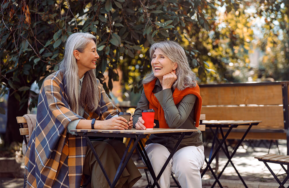 Two senior women sitting at an outdoor table, enjoying coffee and laughing together.
