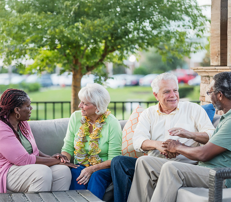 Four seniors enjoying conversation on a patio in a lush garden setting.