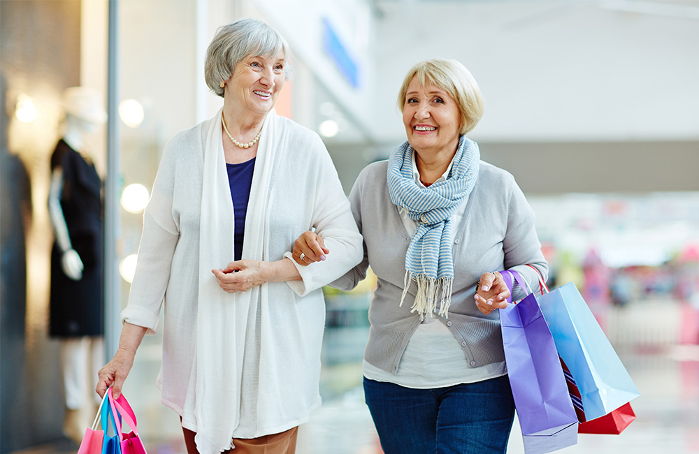 Two senior women smiling and carrying shopping bags in a mall.