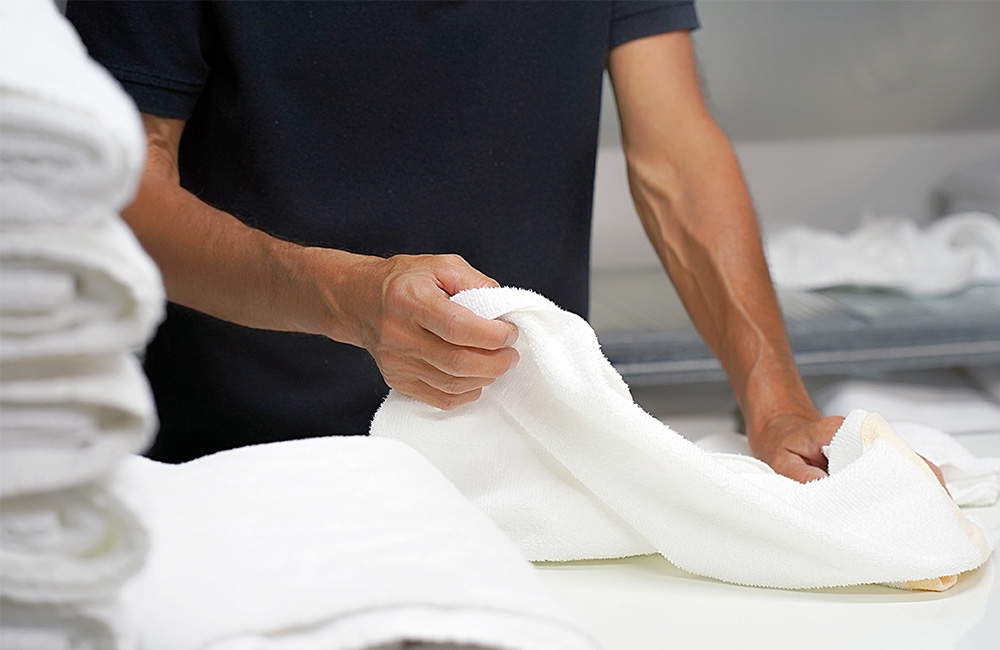 Person folding white towels in a laundry room with stacks of towels nearby.