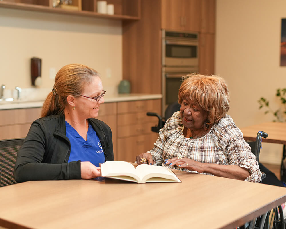 Caregiver assisting a resident with reading in a cozy community space.