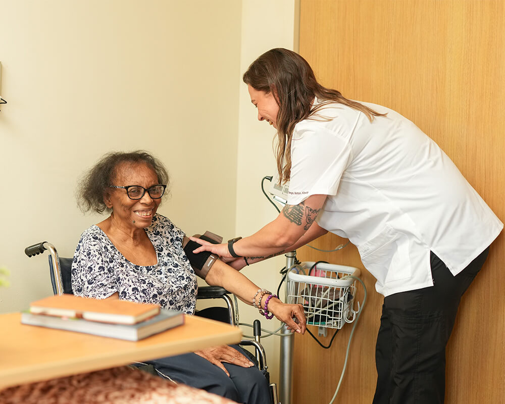 Caregiver assisting an elderly man with exercise in a senior living unit.