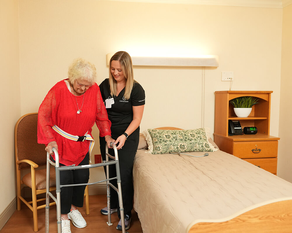 Caregiver assists resident with walker in a cozy unit bedroom.