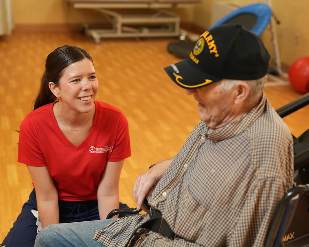Caregiver smiles at elderly man in a wheelchair inside a communal area.