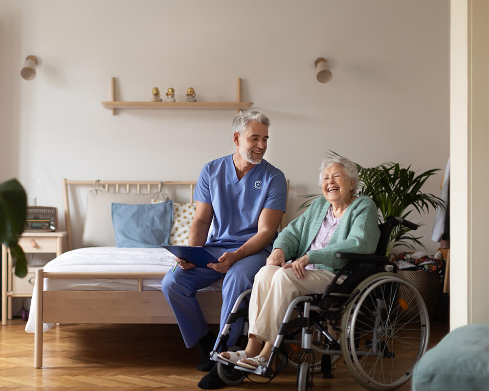 Caregiver in blue scrubs smiling with senior woman in wheelchair in a cozy living unit.
